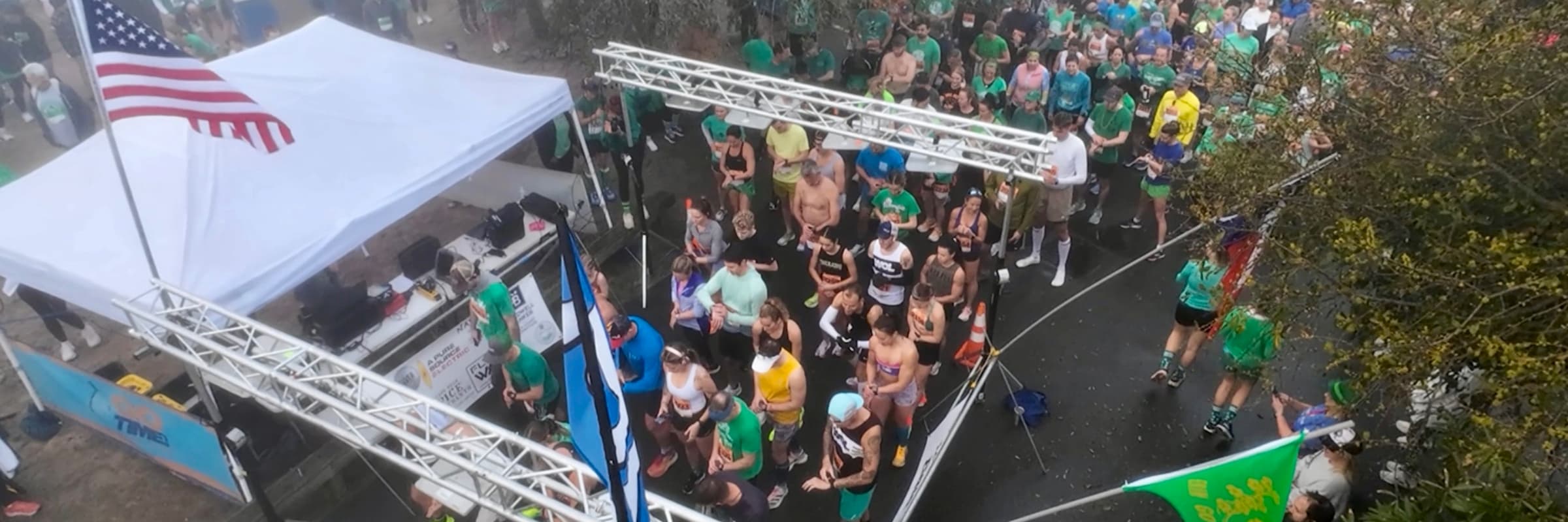 Overhead view of runners gathering at the Lo-Tide Run start line beside a tent, race truss, and American flag.